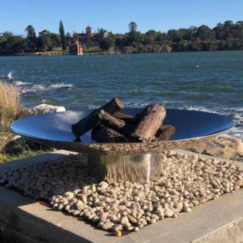 A stainless steel fire pit with logs ready to burn, placed on a rocky base with a reflective surface, overlooking a body of water.