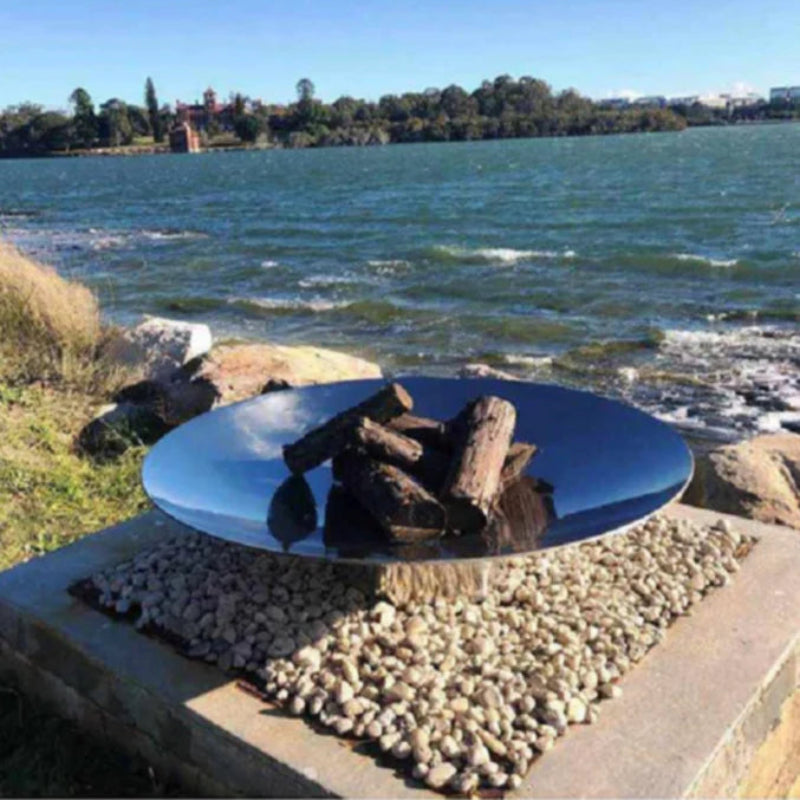 A stainless steel fire pit with logs ready to burn, placed on a rocky base with a reflective surface, besides water with trees in the background.