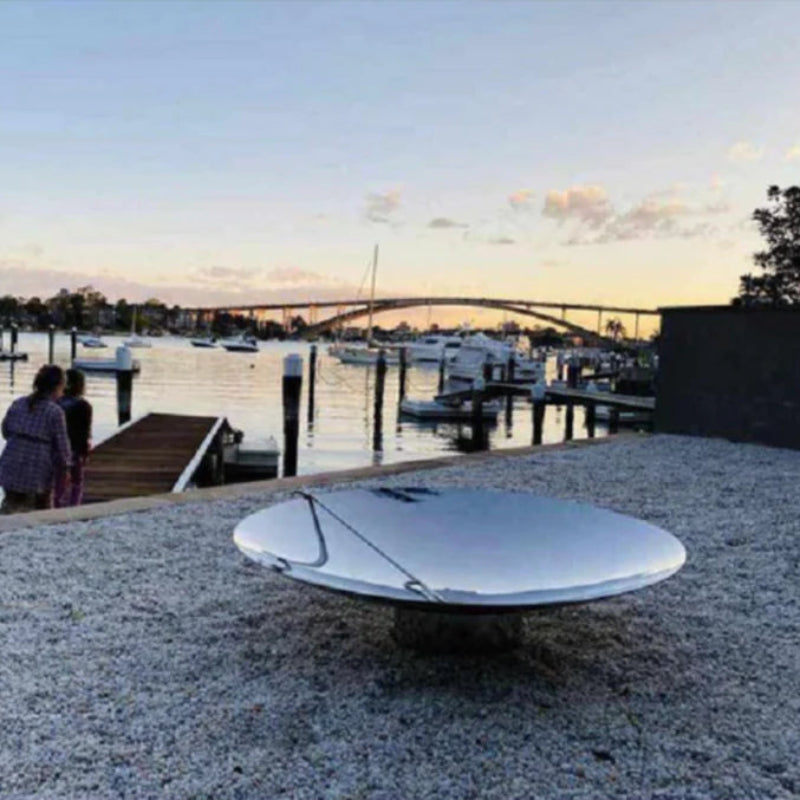 A stainless steel fire pit with poker, placed on a gravel ground with a reflective surface, besides body of water with jetty, boats and a bridge in the background.