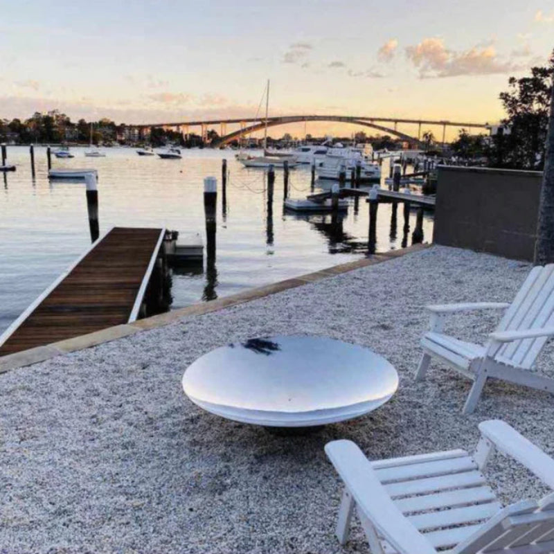 A stainless steel fire pit on a gravel ground with a reflective surface, and a couple of wooden deck chairs besides a body of water with jetty, boats and a bridge in the background.