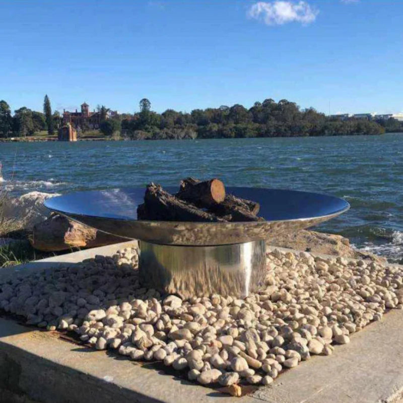 A stainless steel fire pit with logs ready to burn, placed on a rocky base with a reflective surface, overlooking a body of water.