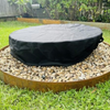 Large round firepit with vinyl cover on, sitting in a gravel fire pit area with grass around it, fence and small trees in the background.
