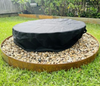 A fire pit with vinyl cover in a backyard setting on a pebble surrounding with fence and garden in the background.