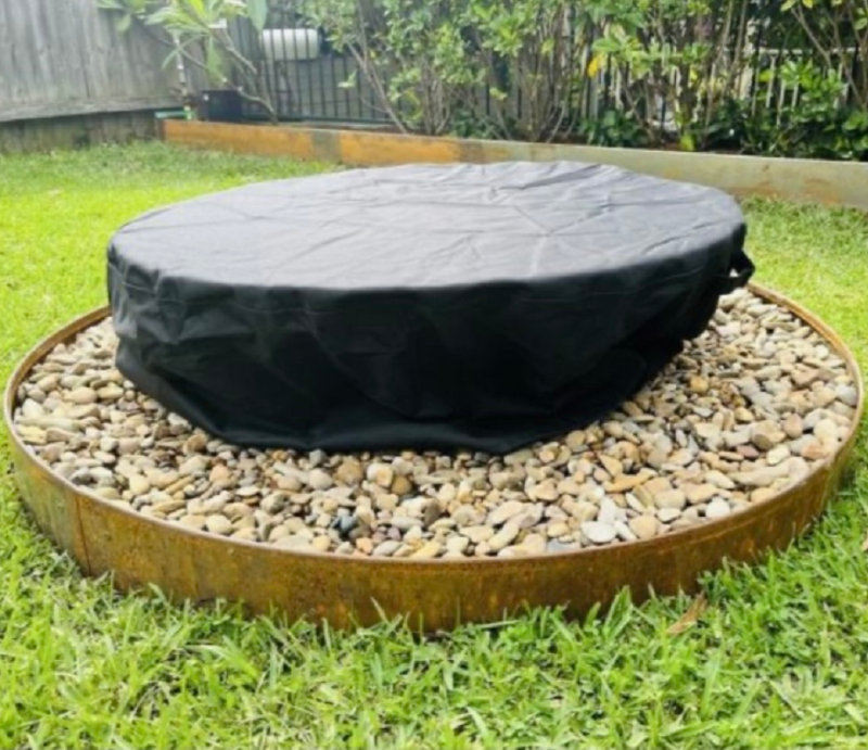 A fire pit with vinyl cover in a backyard setting on a pebble surrounding with fence and garden in the background.