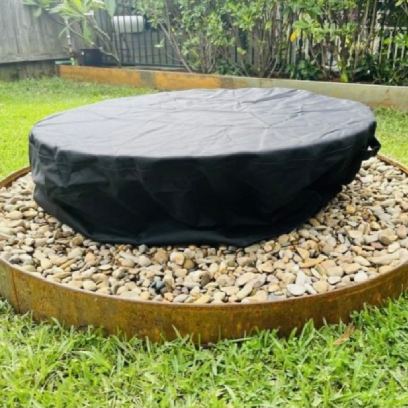 A round shaped fire pit with vinyl cover sitting on a gravel surround with a lawned area and plants in the background.