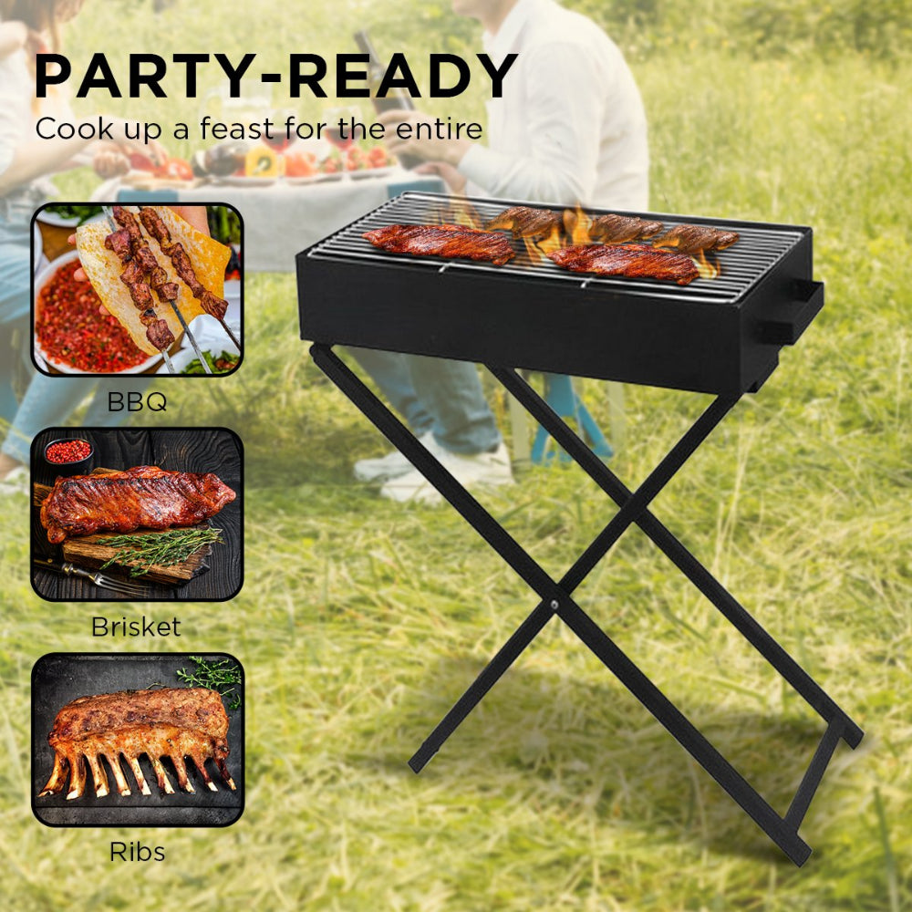 A portable charcoal grill with meat cooking and foldable legs on a stand in a garden setting with a couple sitting and eating at a table in the background.