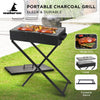 A portable charcoal grill with meat cooking and foldable legs on a stand in a garden setting next to a patio area.