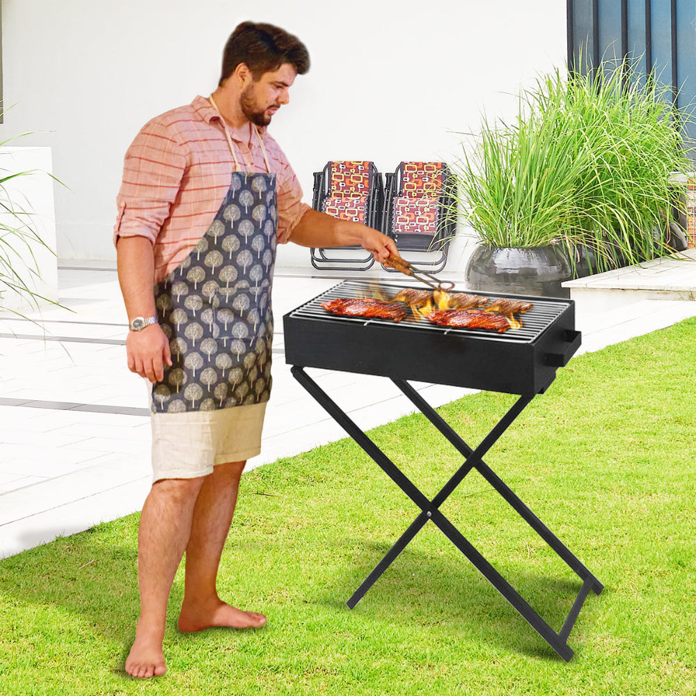 Man cooking at a portable charcoal grill with foldable legs on a stand in a lawn garden setting with patio beside it.