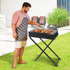 Man cooking at a portable charcoal grill with foldable legs on a stand in a lawn garden setting with patio beside it.