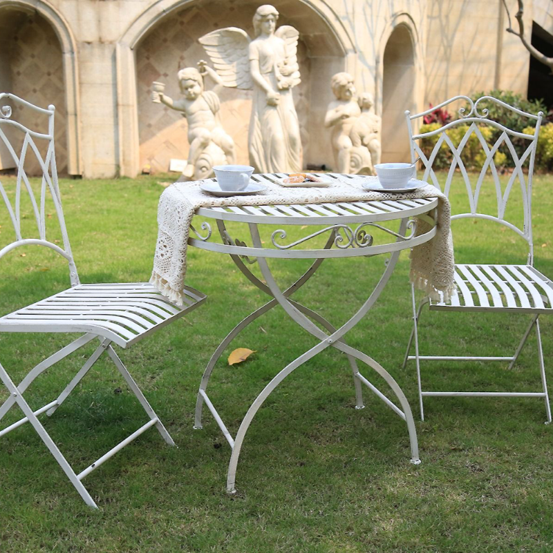 White coloured Albany 3 Piece Wrought Iron Setting in a garden setting with a tablecloth, 2 cups and saucers and plate of biscuits on the table. On a green lawn with marble building, statues and bushes in the background.