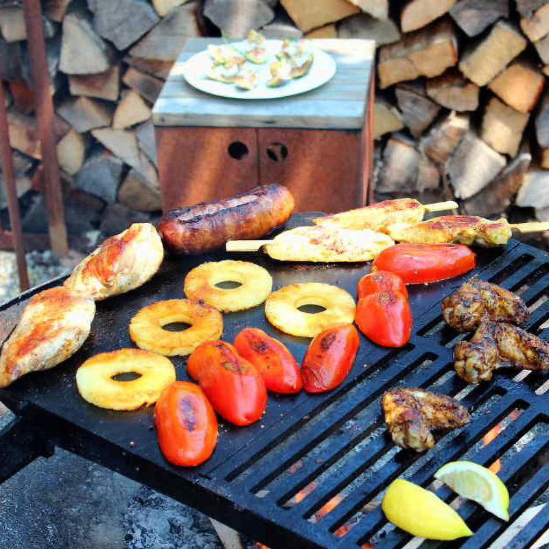 Yagoona Yabbi Fire Pit and Barramundi BBQ Australia with sausages, chicken pieces, tomatoe halves, pineapple rings and lemon wedges. Logs stacked in the background and a side table with stuffed figs on.