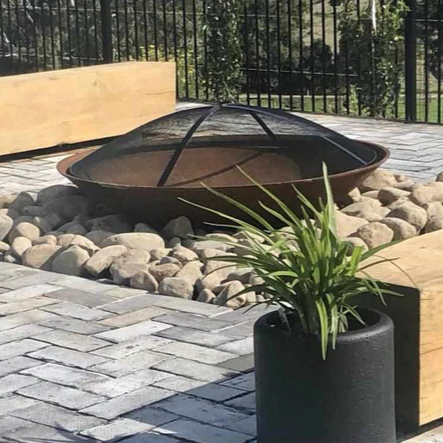 A circular metal ember screen on a round fire pit, featuring in a garden setting with rocks, paving and wooden seating blocks.