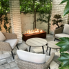Corten coloured Galio Linear Automatic Gas Fire Pit on a tiled patio with wicker furniture, a couple of wooden coffee tables, bonsai tree and other garden trees close by.