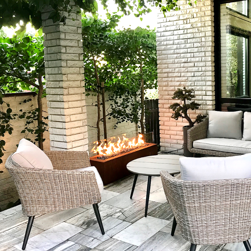 Private residence with Galio Linear Automatic Gas Fire Pit in Corten colour on a tiled patio with wicker furniture, a bonsai tree and several garden trees close by.