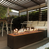 A brown, rectangular-shaped outdoor gas fire pit with a protective glass fender and decorative black diamond stones. It is placed on a wooden deck with plants in the background.