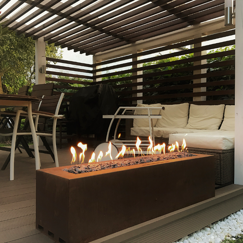 A brown, rectangular-shaped outdoor gas fire pit with a protective glass fender and decorative black diamond stones. It is placed on a wooden deck with plants in the background.