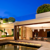 Galio Gas Fire Pit Insert Linear Automatic along the front of a swimming pool with wood decking, a burning fire in front of a house with open glass doors into a dining room with table and 8 chairs, large round shaped bushes in big planters on the roof top and large pot with plants around the side of the house, and a beautiful blue sky.