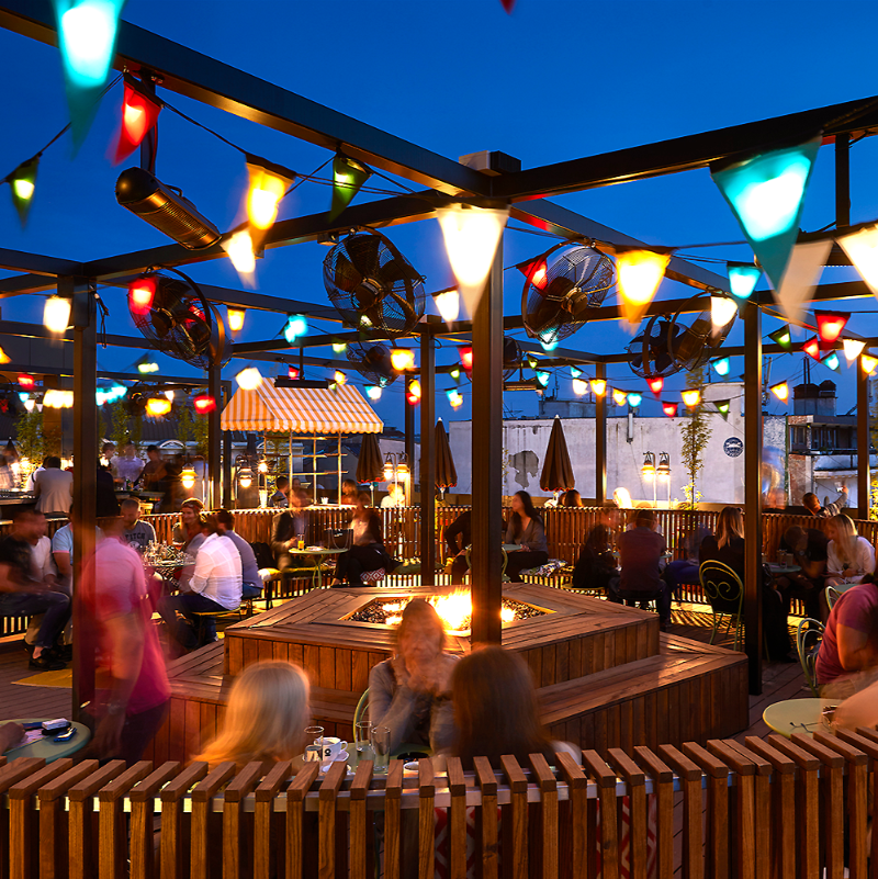 Galio Gas Fire Pit Insert Linear Automatic in an outdoor entertainment area, looks like a pub, lots of people sitting around, fans and flags set up on large pergola, wooden decking and seating all around the place.