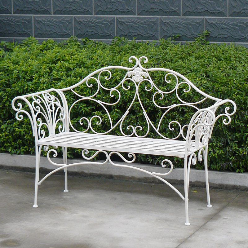 Katerina Antique white Iron Bench in front of a green hedge, grey brick wall and on a cement flooring.