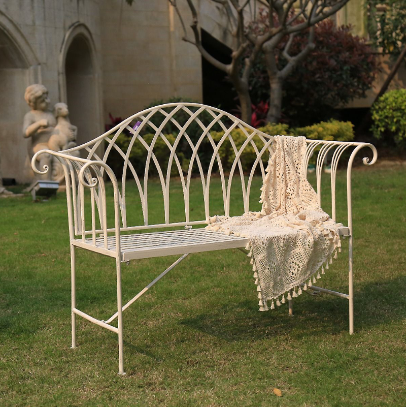 Lavinia Iron Bench Australia Antique White in garden setting with a cream coloured croqueted throw on green grass with a building, shrubs, trees and cement statue in the background.