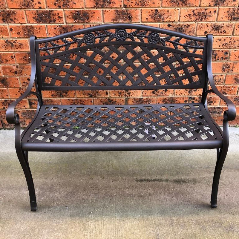 Dark bronze coloured Melissa Cast Aluminium Bench on a cemented floor area in front of a brown and orange brick wall.