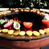 Food cooking on Yagoona Ringgrill BBQ & Goanna Fire Pit, fire can be seen in the centre of the firepit, bushes in the background.
