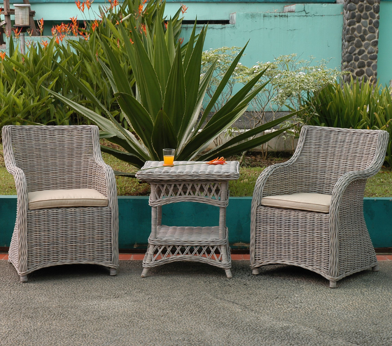 Roma 3 Piece Natural Aged KUBU Wicker Set in an outdoor setting, a glass of orange juice and a single flower on the tabletop, bushy garden and flowers in the backdrop.