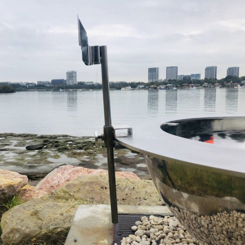 Teppanyaki Spit Rotisserie Bracket  and pole on the side of a fire pit beside a body of water, with buildings and boats in the background.