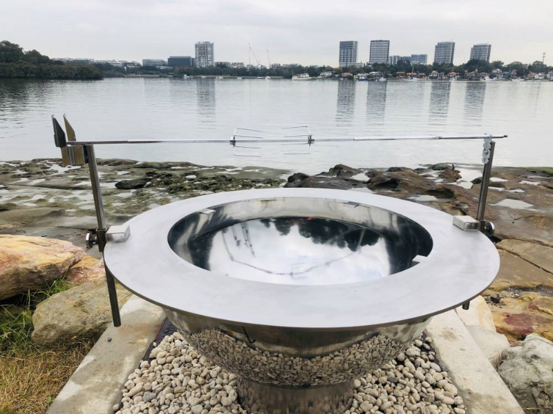Teppanyaki Spit Rotisserie Brackets and poles with rotisserie set up on a fire pit. On a pebble surround next to a body of water, with buildings and boats in the background.