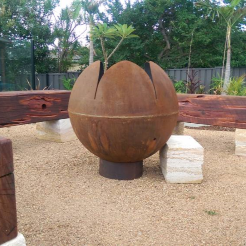 The Rust coloured Lotus shaped Fire Pit in a pretty garden setting, with large wooden logs for sitting on around the fire.