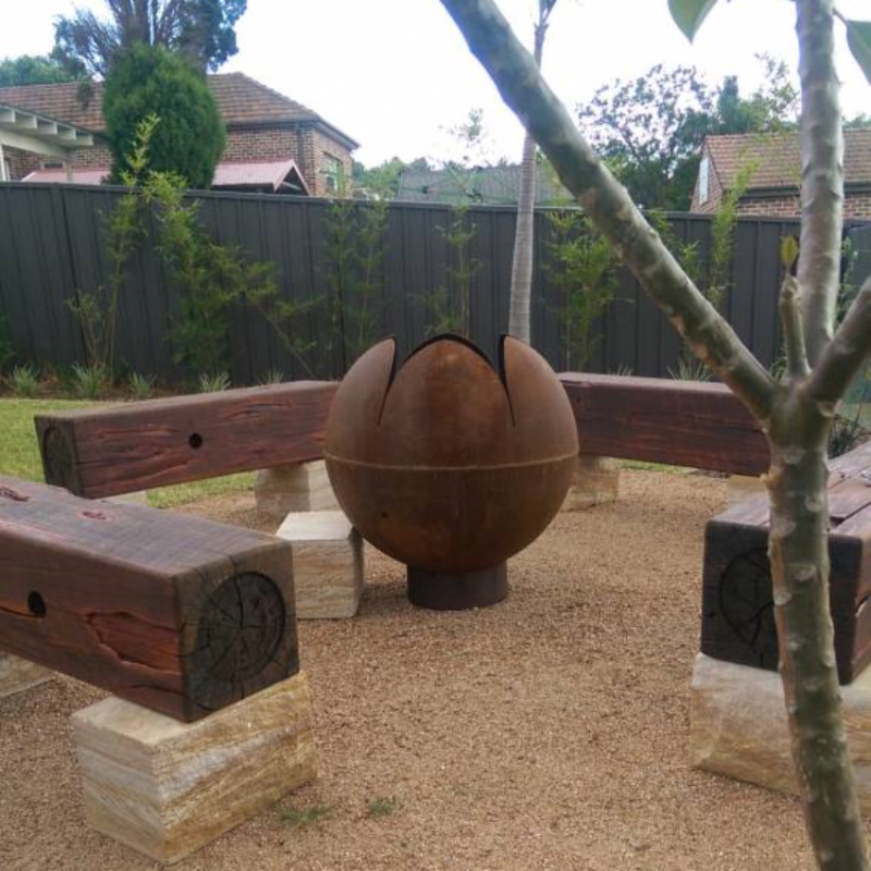 The Lotus Fire Pit in a nice back yard setting, large wooden bench seating around it, fence and trees in the backdrop.
