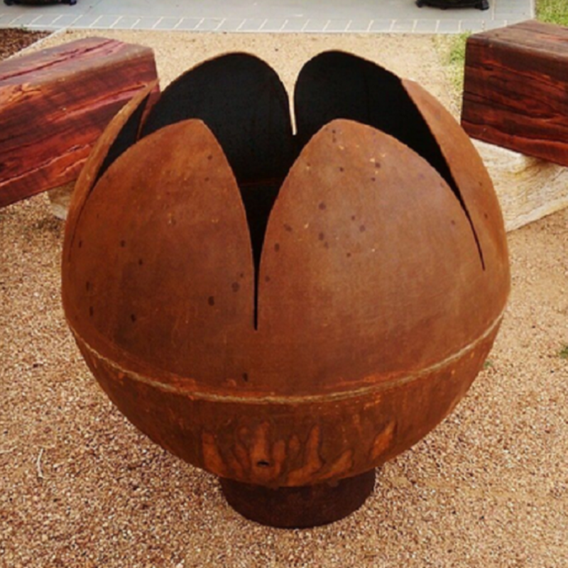 The Lotus Fire Pit in natural rust coloured sitting on a sandy stone surface with big wood logs for seating close by.