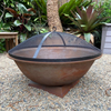 A Rust colour cast iron round basin fire pit on stand with an ember screen on a gravel surface in a garden setting.