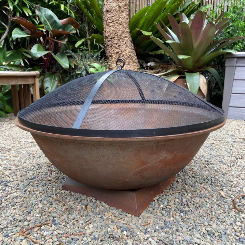 A Rust colour cast iron round basin fire pit on stand with an ember screen on a gravel surface in a garden setting.