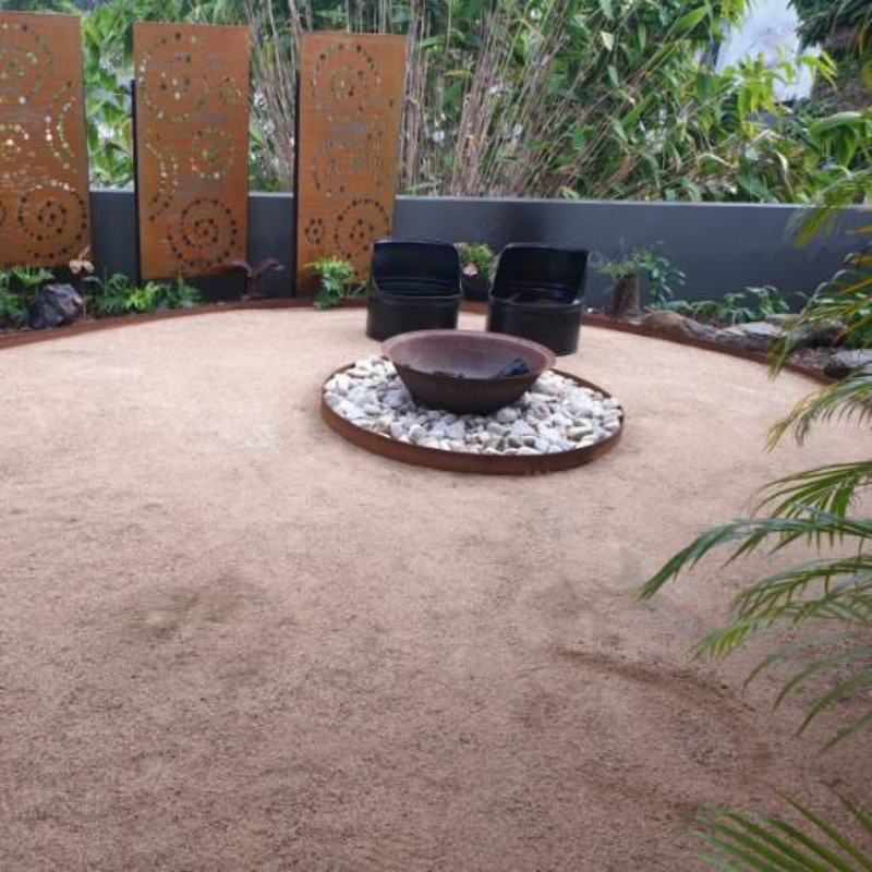 Rust coloured cast iron round basin fire pits with a rock surround and a couple of outdoor seats and private garden setting in the background.