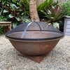 A Rust colour cast iron round basin fire pit on stand with an ember screen on a gravel surface in a garden setting.