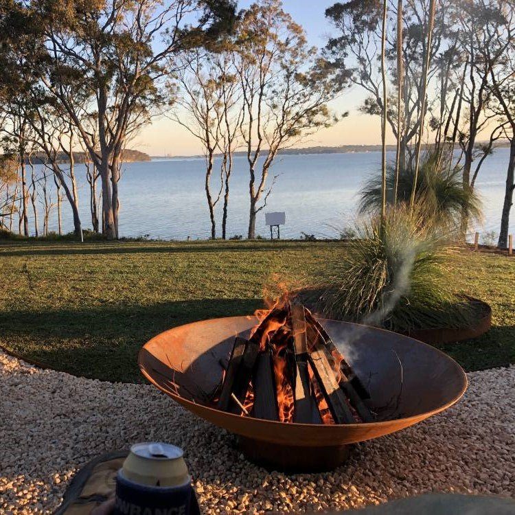 Rust-colored bowl shape metal fire pit with a fire burning near a lawn and trees and a lake in the background.