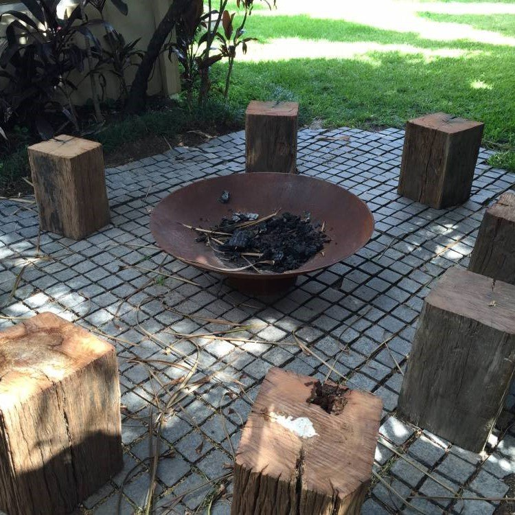 Rust-colored bowl shape metal fire pit with remains of fire near a lawn on a paved area with tree stump chairs.
