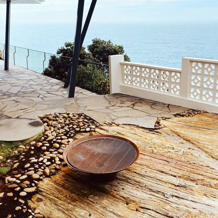 Rust-colored bowl shape metal fire pit with stainless steel grill on a patio area overlooking the ocean.