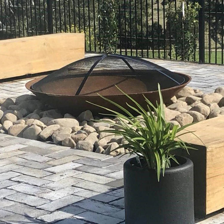 Rust-colored bowl shape metal fire pit with ember screen on a backyard setting with trees and hills in the background.