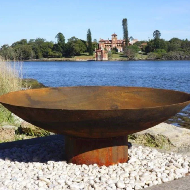 Rust-colored metal bowl shape fire pit on pebbles with a lake and distant building in the background
