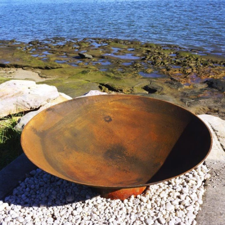 Rust-colored metal bowl fire pit on a pebbled surface with water and rocks in the background