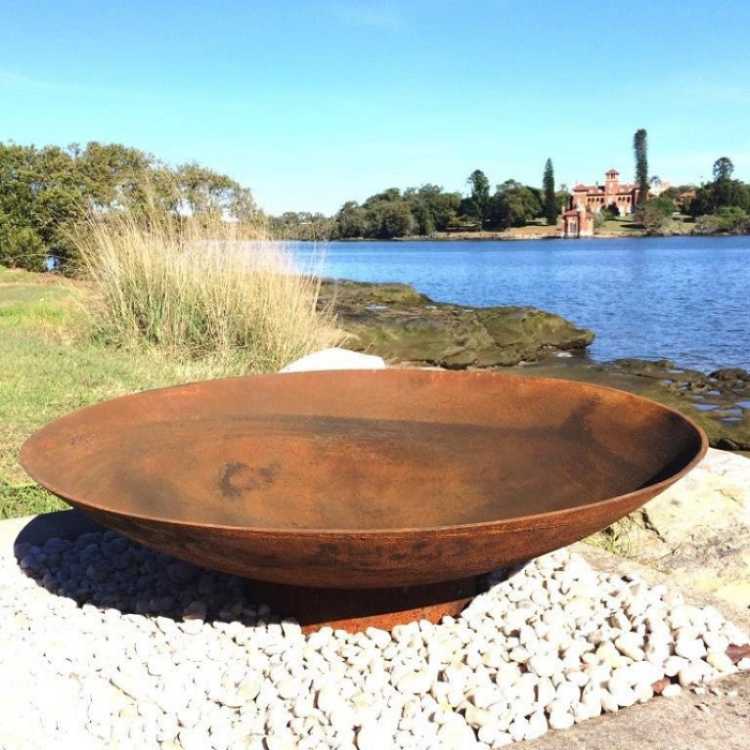 Rust-colored metal fire pit on a pebble surface with a scenic background of water and trees.