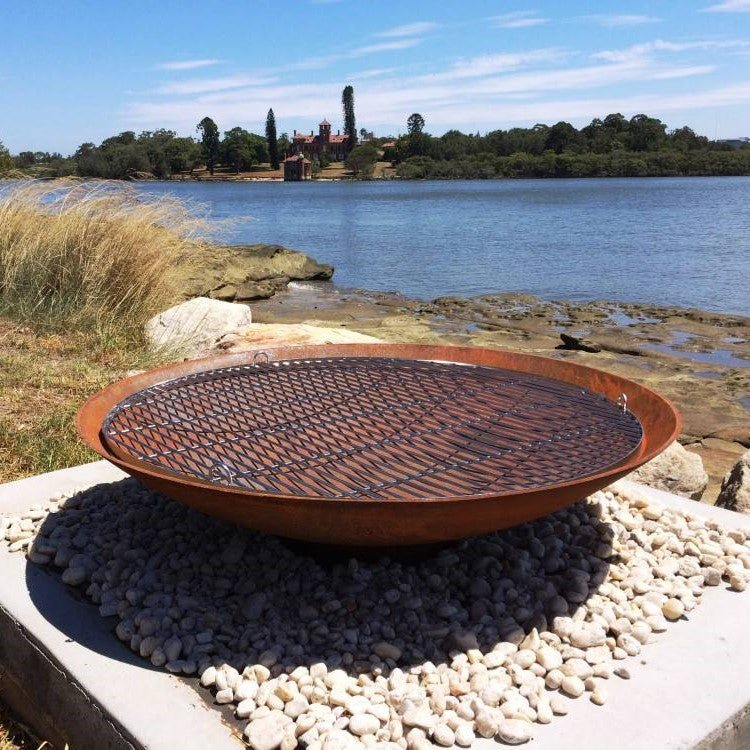 Rust-colored metal fire pit with stainless steel grill on a pebble surface with a scenic background of water and trees.