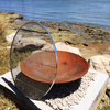 Rust-colored metal fire pit and a stainless steel grill on a pebble surface with a scenic water background.