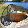 Rust-colored metal fire pit and a metal grill on a pebble surface with a scenic water and trees background.