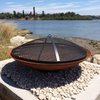 Rust-colored metal fire pit with a 
grill and ember screen on a pebble surface with a scenic water and trees background.
