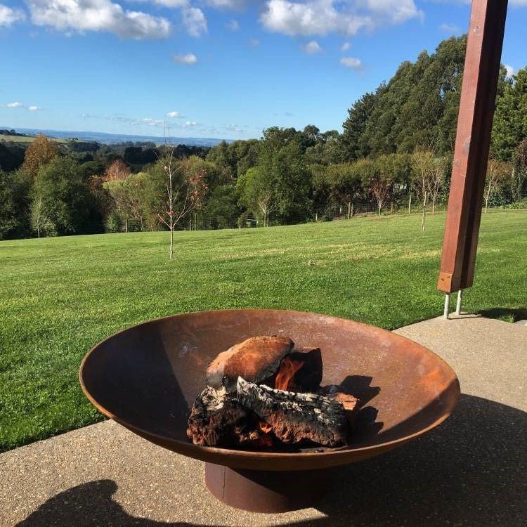 Rust-colored bowl shape metal fire pit with a few burning logs with lawn and scenic trees in the background.