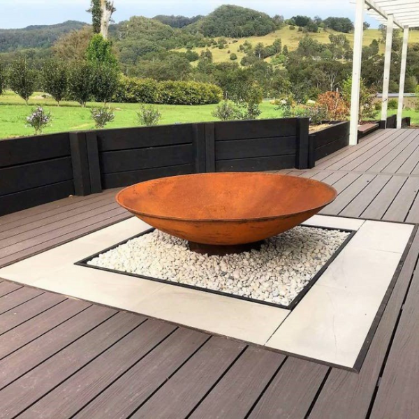 Rust-colored fire pit on a wooden deck with pebble and tiled surround, and scenic background of hills and trees