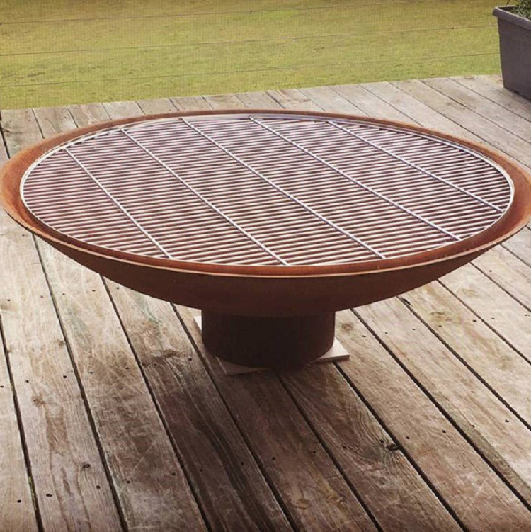 A large round rust colour cast iron fire pit, with stainless steel grill on it placed on a wood deck with lawn area next to it.